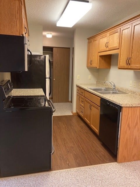 A narrow kitchen with wood cabinets and black appliances, featuring a stove, refrigerator, sink, and dishwasher.