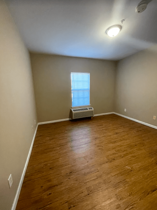 An empty room with laminate flooring and a single window, featuring neutral-colored walls and a ceiling light.