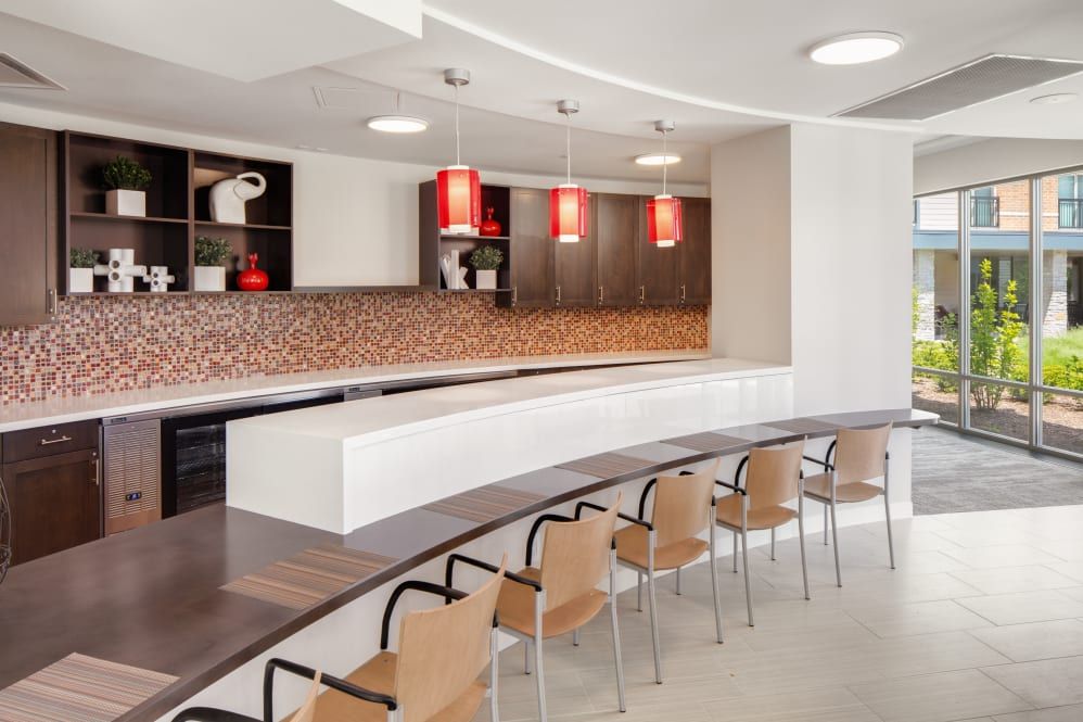 A modern kitchen interior with a long countertop, pendant lights, and rows of chairs.