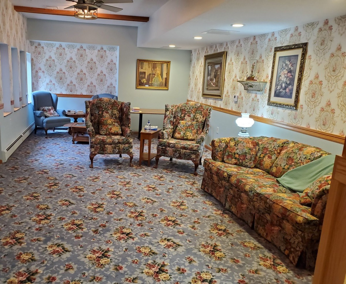 A cozy living room with floral patterned furniture and carpet, featuring armchairs, a sofa, decorative wallpaper, paintings, and a ceiling fan.