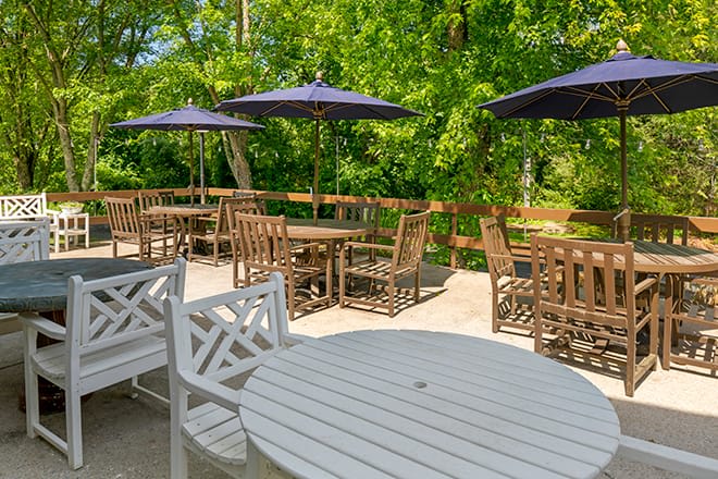 A peaceful outdoor patio area with wooden tables, chairs, and umbrellas surrounded by lush greenery.