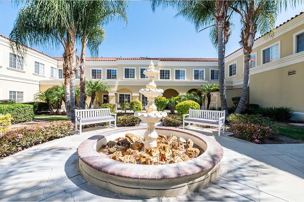 A beautiful courtyard with a central fountain, surrounded by benches and palm trees in front of a building.