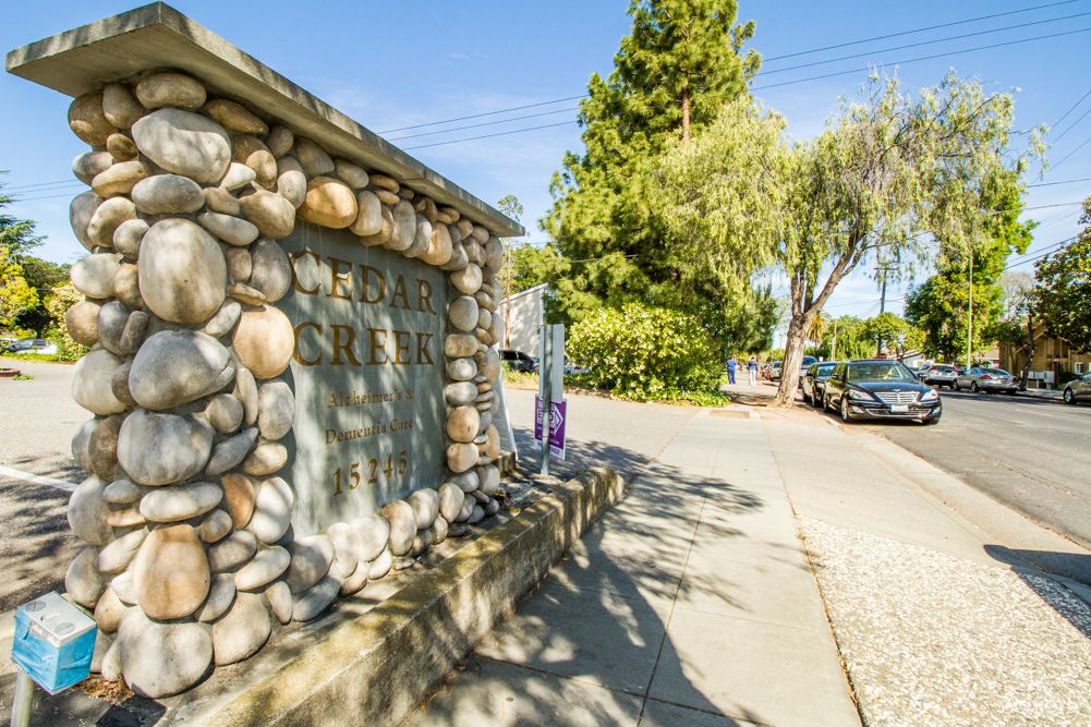 A stone wall sign by a road with trees and parked cars in the background on a sunny day.
