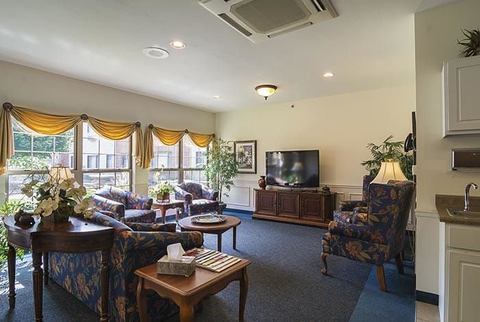 A cozy living room featuring comfortable seating, a television, and natural light streaming through large windows with draped curtains.