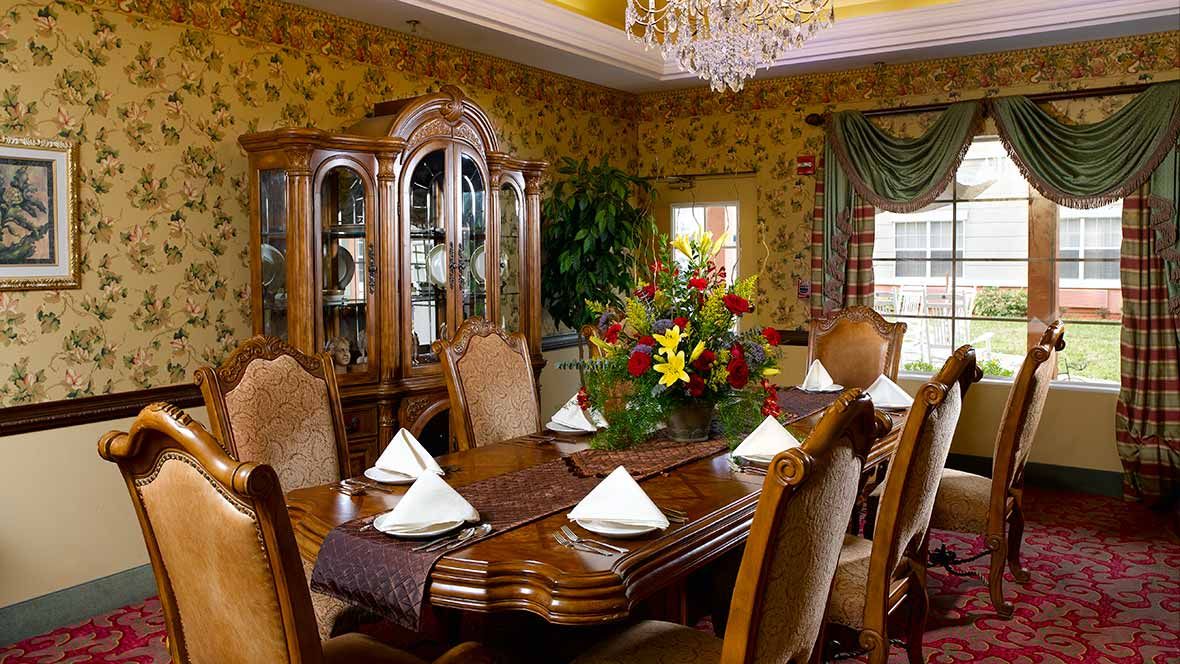 An elegant dining room with a large wooden table, ornate chairs, and a chandelier overhead. A floral arrangement serves as the centerpiece on the table.