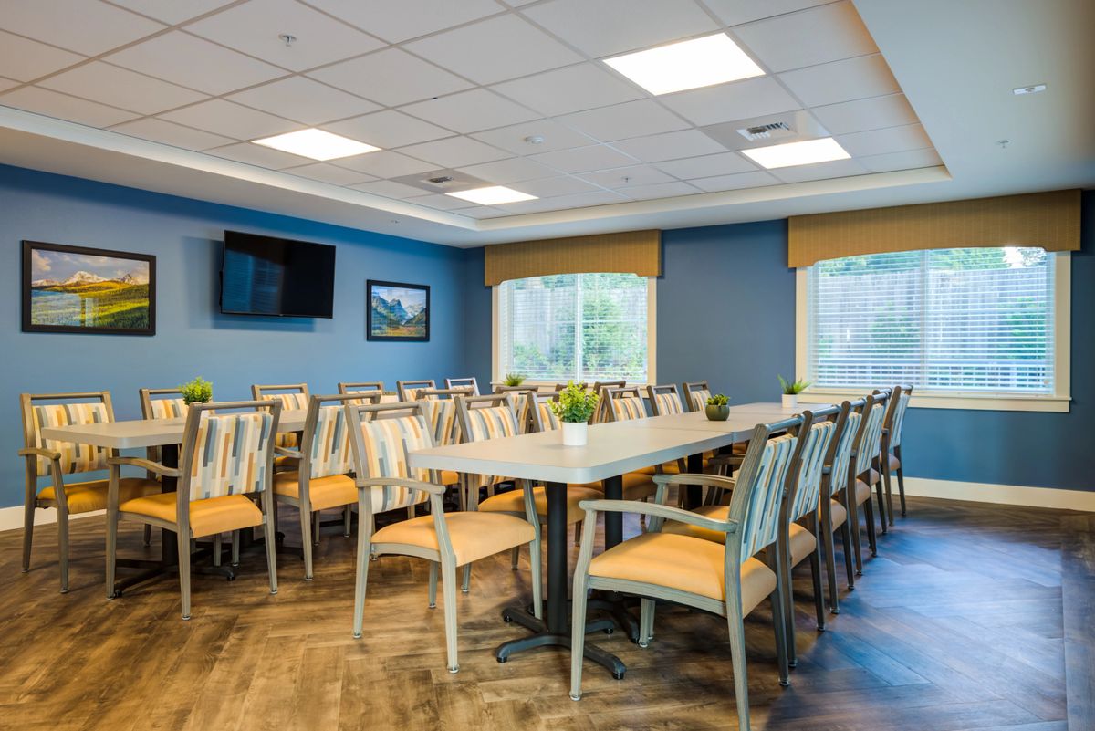 A spacious dining room with arranged tables and chairs, featuring a television on the wall and large windows.