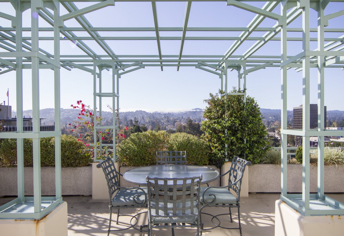 A picturesque outdoor patio with a pergola, set with a table and chairs, surrounded by greenery and offering a scenic view.