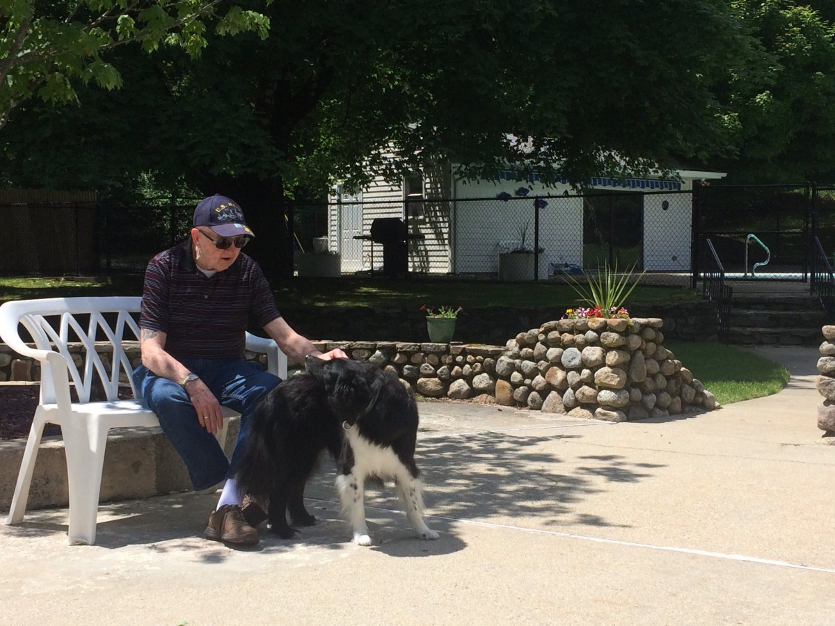 A man sits outdoors petting a dog under a sunny sky.