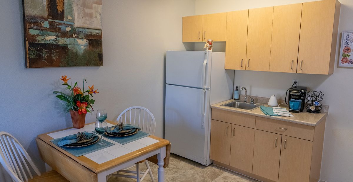 A cozy kitchen and dining area featuring a refrigerator, sink, cabinets, and a neatly set table for two.