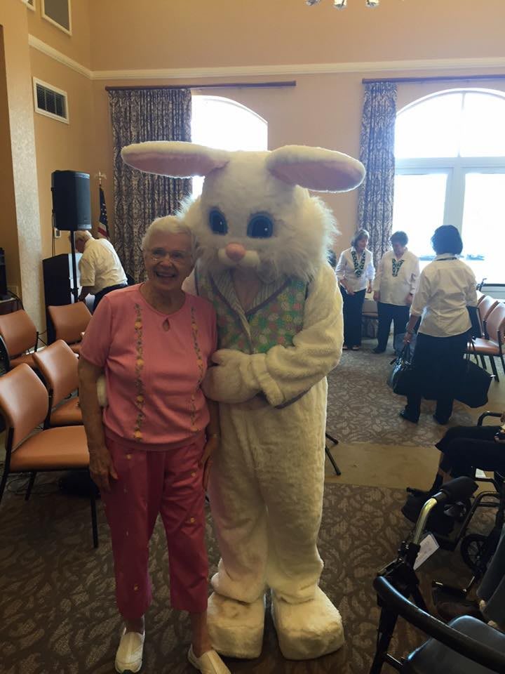 An elderly woman poses with a person in an Easter bunny costume during a celebration indoors.