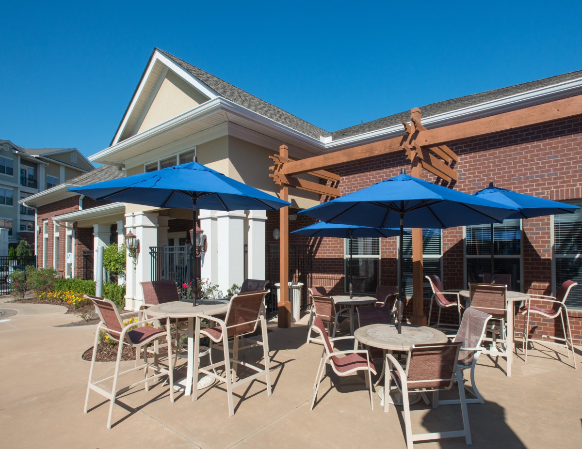 An outdoor patio area with tables and chairs shaded by blue umbrellas, set against the backdrop of a building on a sunny day.