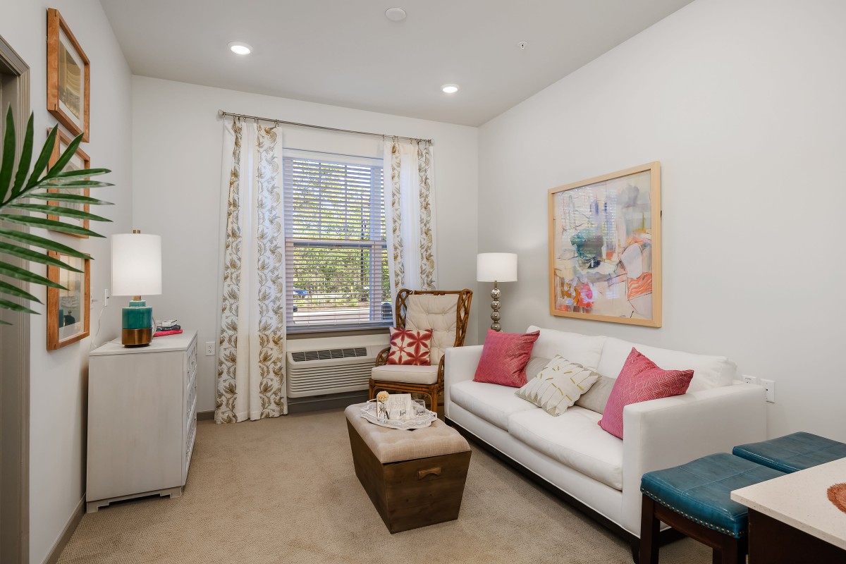 A cozy living room with a white sofa and colorful cushions, complemented by an armchair, decorative curtains, and a framed artwork on the wall.