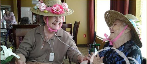 An elderly woman wearing a floral hat enjoys a cheerful moment, radiating joy while interacting with another person.