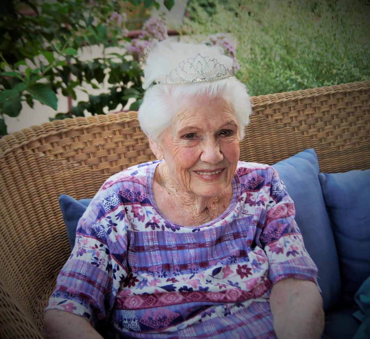 An elderly woman sitting comfortably on a wicker chair with blue cushions, enjoying the garden ambiance.