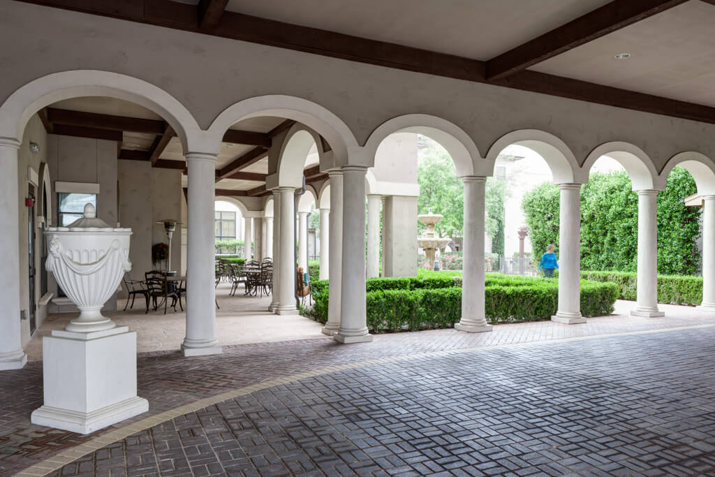 A serene courtyard with archways and white columns, featuring greenery and outdoor seating.