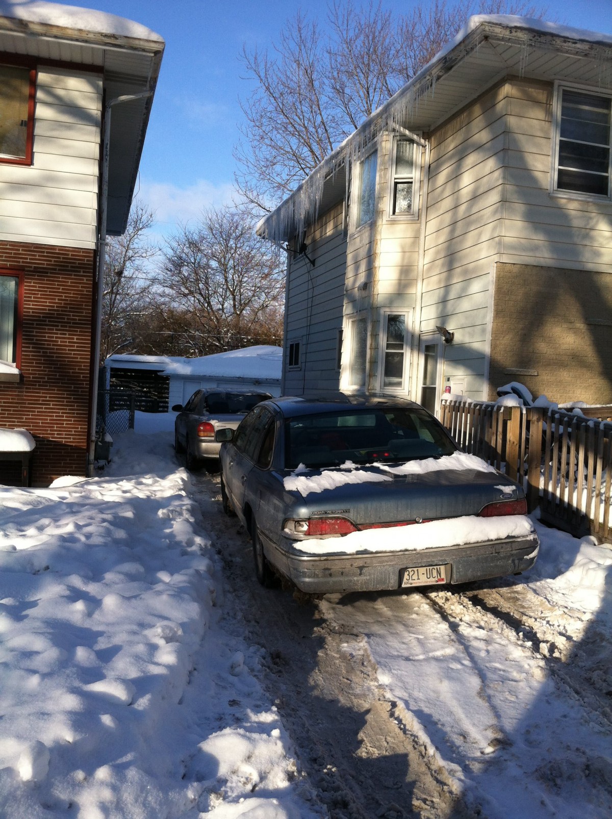 Cars parked in a snowy driveway between two houses on a sunny winter day.