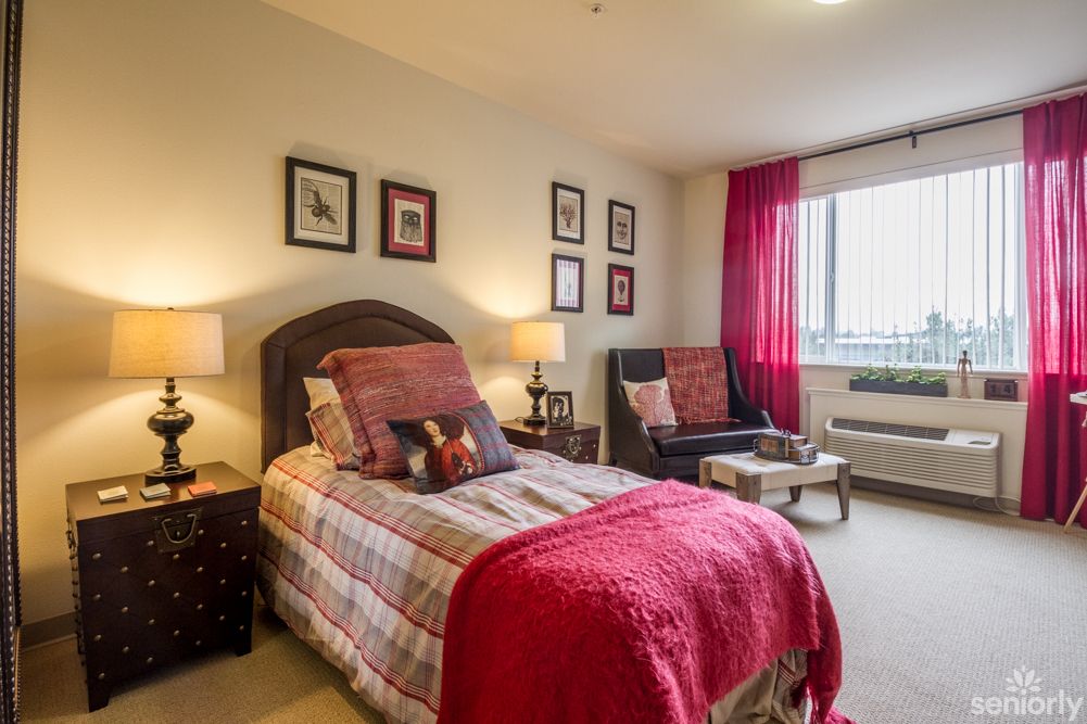 A cozy bedroom with a single bed, adorned with decorative pillows and warm lighting, featuring framed artwork on the walls and a comfortable armchair by the window.