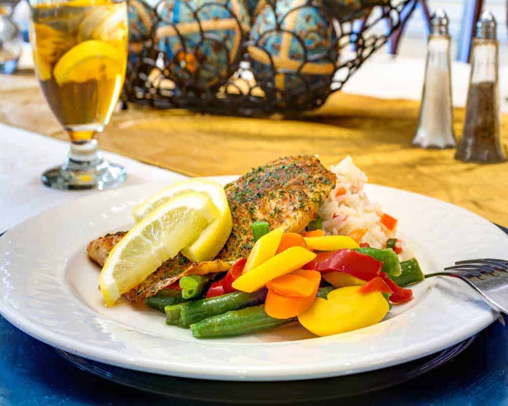 A plate of seasoned fish with colorful vegetables and lemon slices, accompanied by a glass of iced tea.