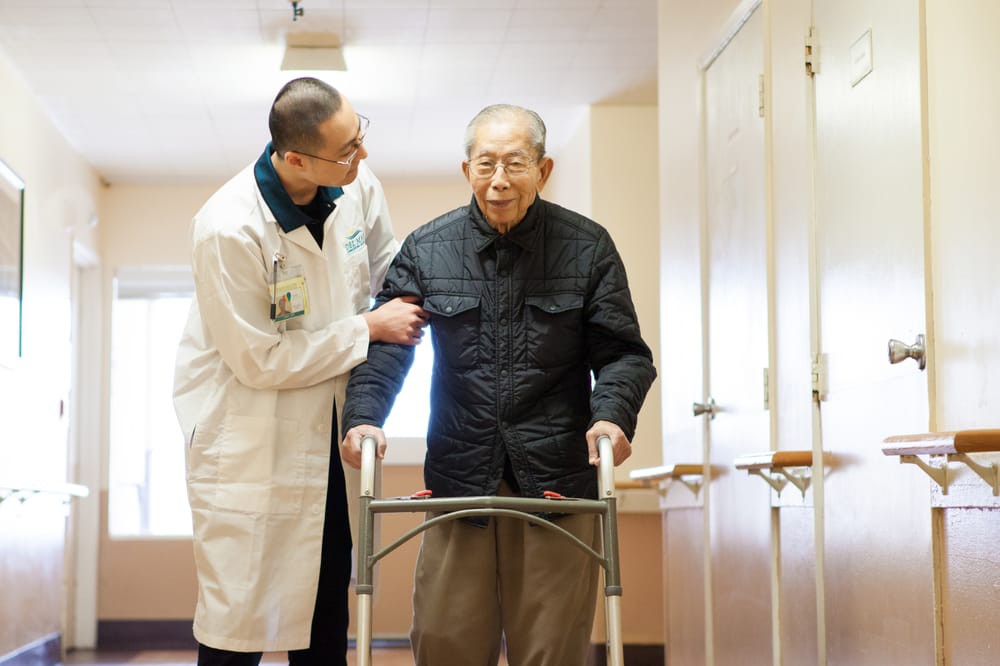 A doctor assists an elderly man using a walker in a hallway.