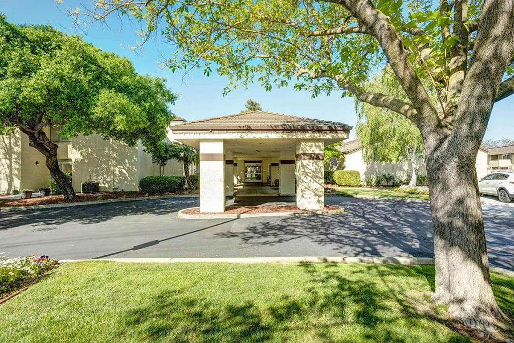 A well-maintained building entrance with a driveway, surrounded by trees and greenery on a sunny day.