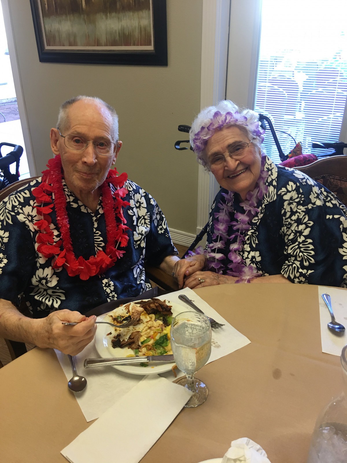 An elderly couple enjoying a meal dressed in tropical-themed outfits with leis around their necks.
