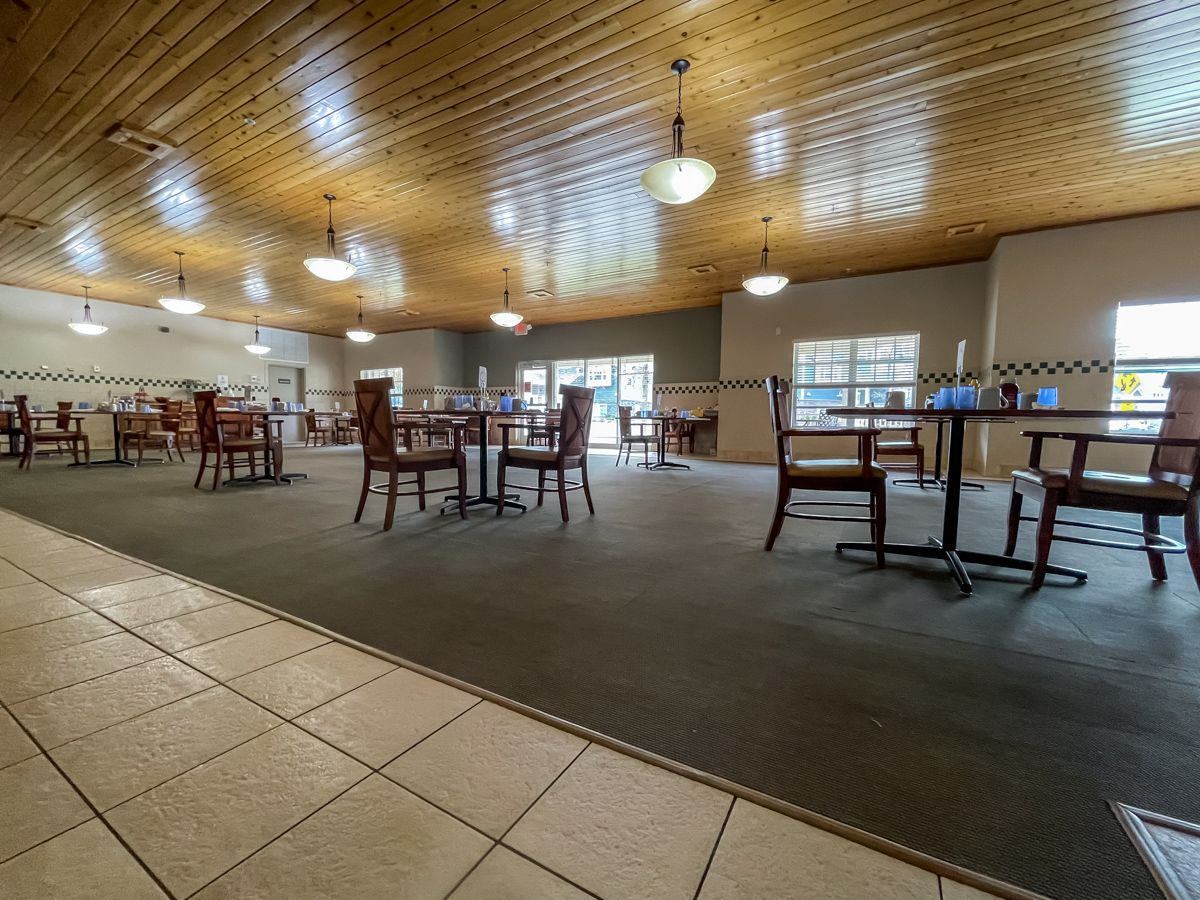 A spacious restaurant dining area with wooden ceilings and a carpeted floor, featuring tables and chairs under ceiling lights.