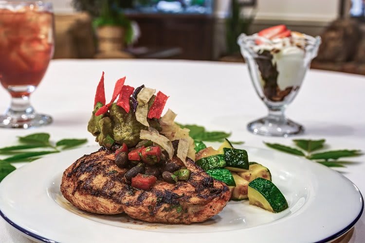 A beautifully plated grilled chicken breast served with sauted vegetables and a dessert in the background.