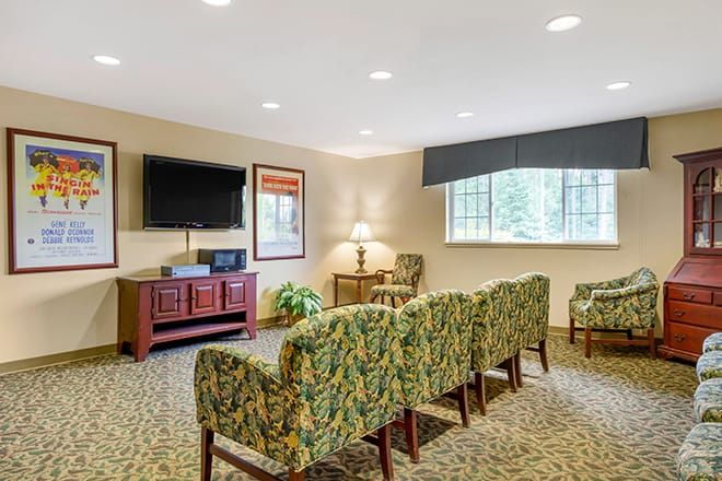 A comfortable waiting room with patterned chairs facing a television, featuring a lamp and artwork on the walls.