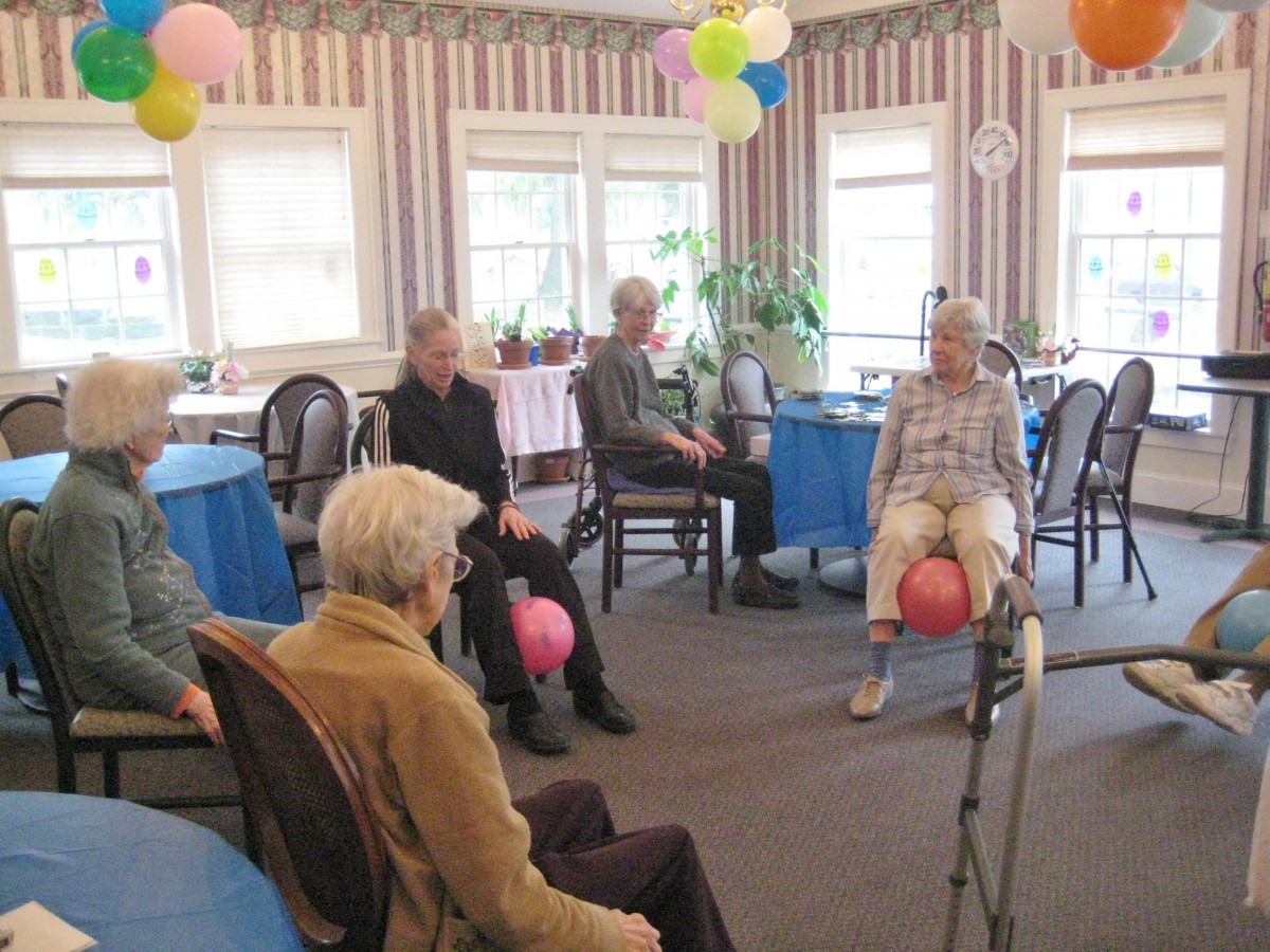 A group of seniors participating in a seated exercise activity with balloons in a community room.