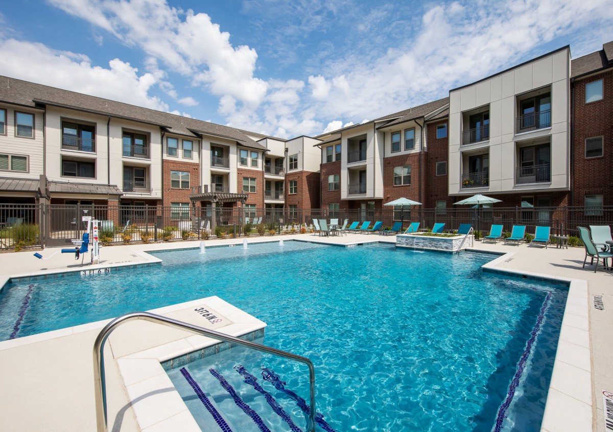 A modern apartment complex featuring a spacious swimming pool and outdoor seating on a sunny day.