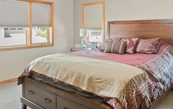 A cozy bedroom with a neatly made bed, featuring decorative pillows and two windows allowing natural light.