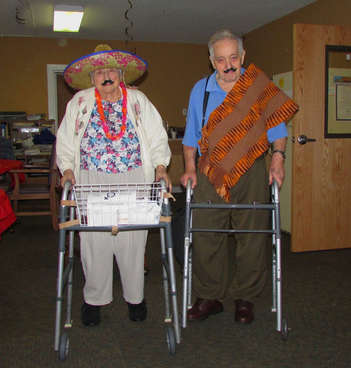 Two elderly individuals dressed in humorous costumes with walkers indoors.