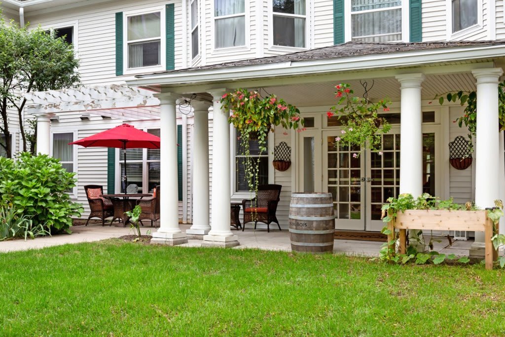 A charming patio with white columns and outdoor seating, featuring a red umbrella and a wooden barrel, set against a lush green lawn.