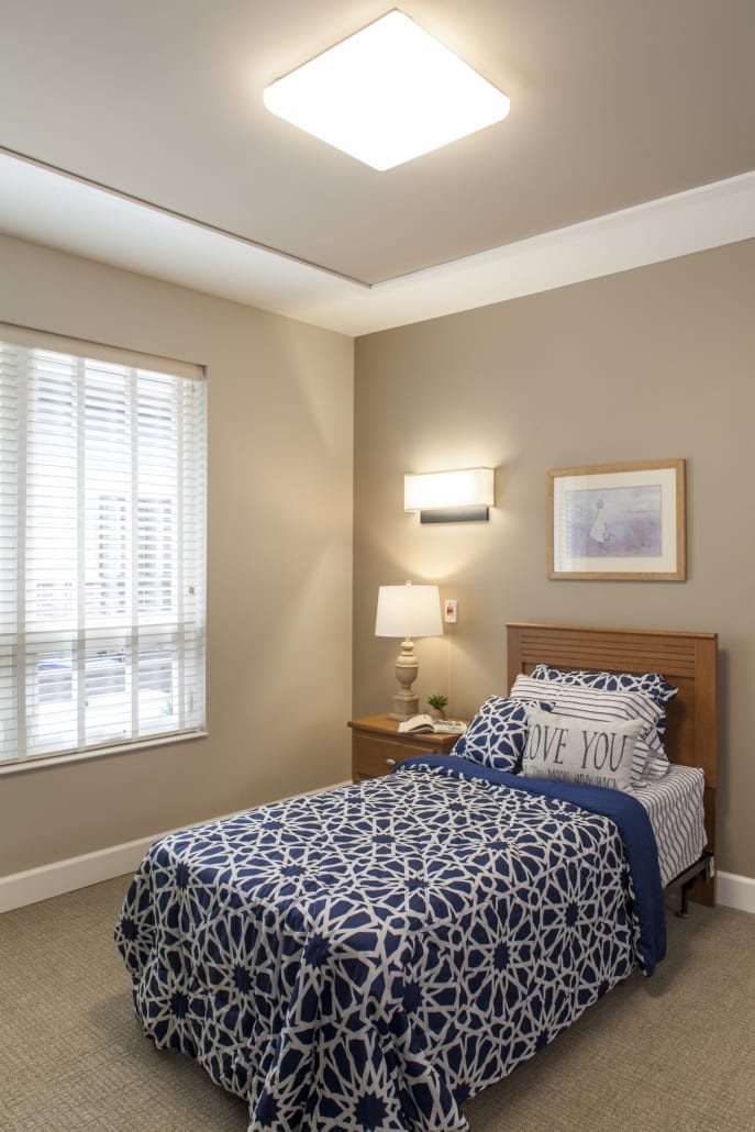 A cozy bedroom with a single bed, decorated in blue and white tones, featuring a window, lamp, and wall art.