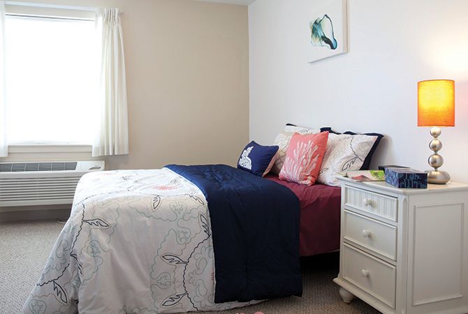 A cozy bedroom with a neatly made bed, featuring colorful pillows and a nightstand by the window.
