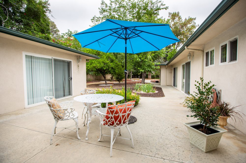 A cozy patio area with a blue umbrella, white metal table, and colorful chairs, surrounded by greenery.