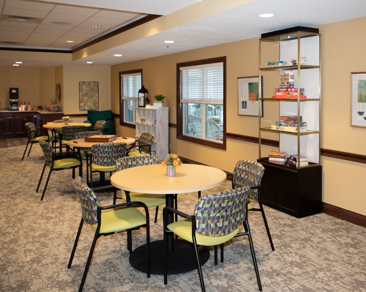 A cozy dining room with round tables and patterned chairs, featuring decorative shelves and large windows.