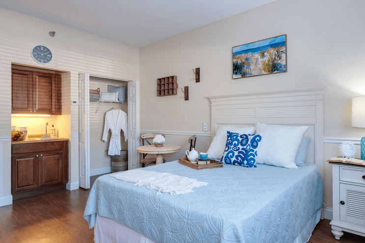 A cozy bedroom with a neatly made bed, featuring blue accents and decorative elements, including a painting and a robe hanging in an open closet.