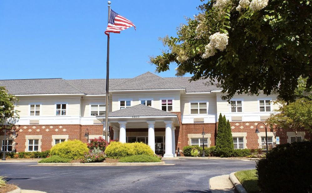 A residential building with an American flag flying in front, surrounded by landscaping and trees under a clear blue sky.