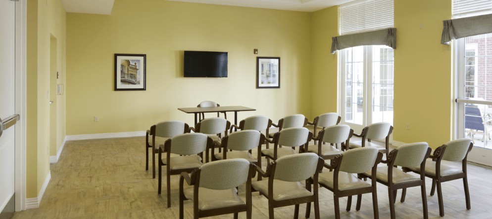 A neatly arranged conference room with rows of chairs facing a table and television, set against yellow walls.