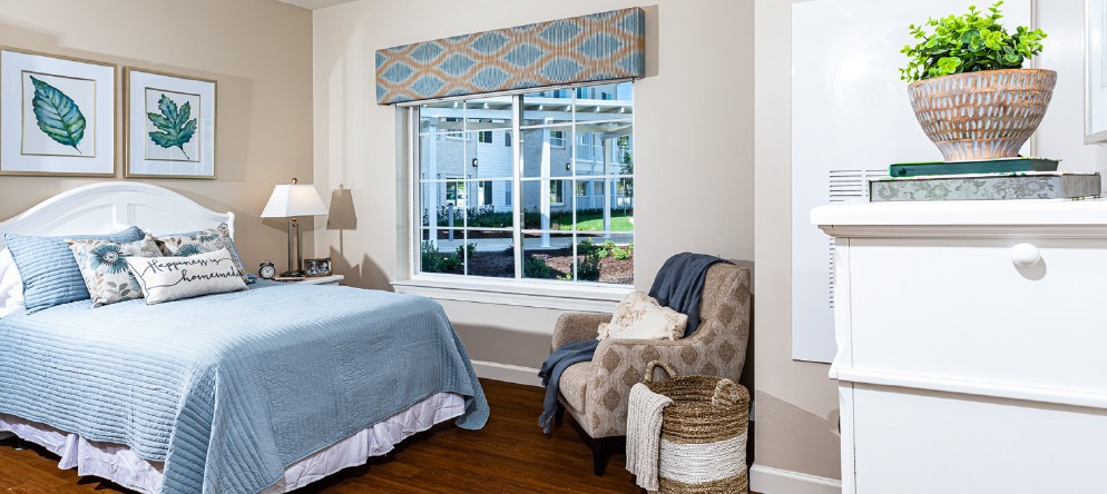 A cozy bedroom with a large window, featuring a blue bedspread, an armchair with a blanket and basket, and a stylish lamp.