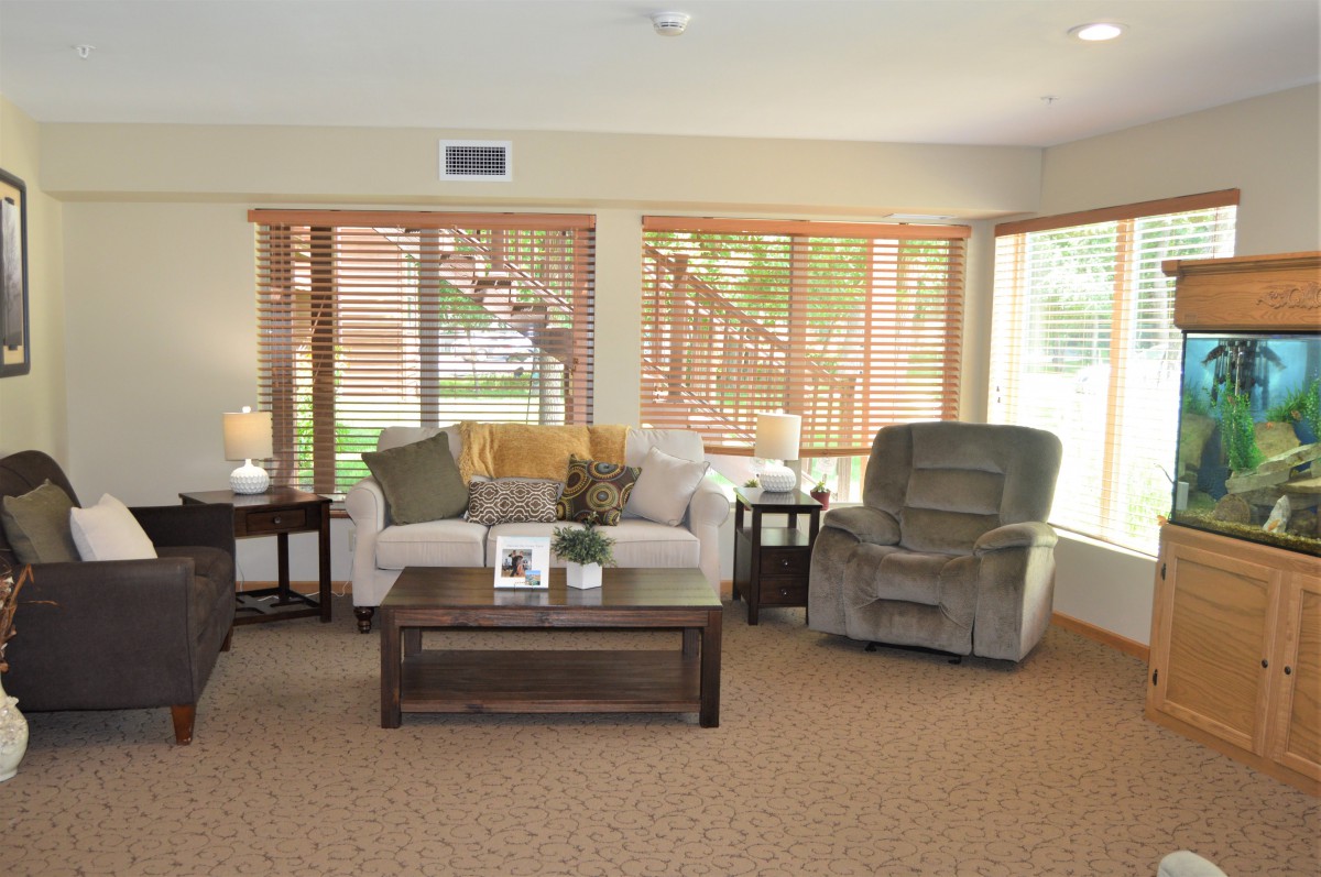 A cozy living room with a sofa, armchair, and coffee table by large windows with blinds.