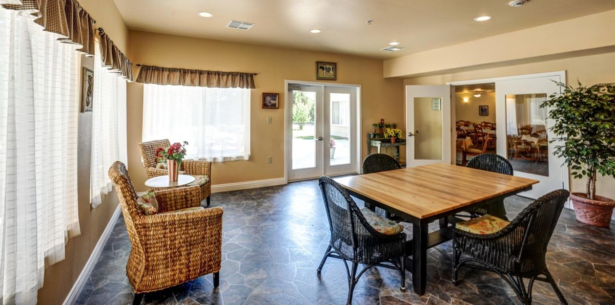 A cozy living room interior with wicker chairs, a wooden table, and ample natural light from large windows.