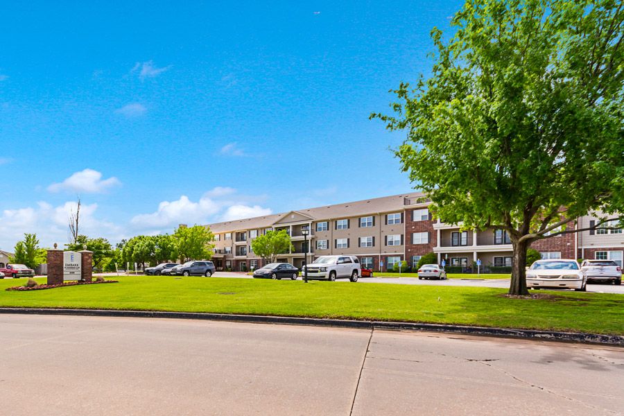 A view of a residential apartment building with a parking lot and surrounding greenery on a sunny day.