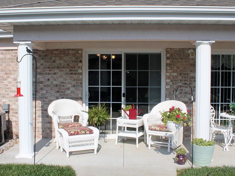 A cozy patio setup with two white chairs and a small table, adorned with plants against a brick wall.