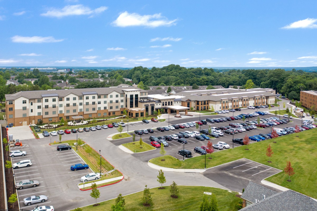 Aerial view of a large building complex with an adjacent parking lot filled with cars, surrounded by greenery and trees under a blue sky.