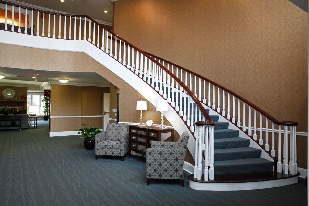 A stylish lobby featuring a sweeping staircase with wooden railing, flanked by two patterned chairs and a table lamp.