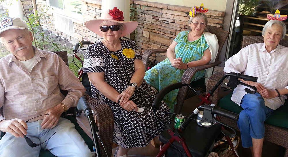 Two elderly women wearing hats and sunglasses sit outside on a patio, each with a walker.