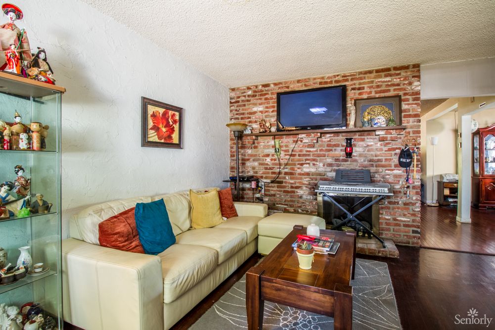 A cozy living room features a beige sectional sofa with colorful cushions, a brick accent wall with a mounted TV, and a keyboard setup beside a wooden coffee table.