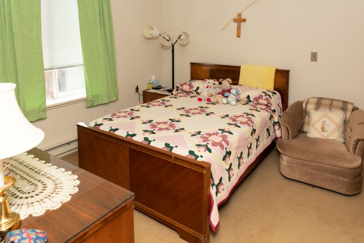 A cozy bedroom with a floral quilt, lamp, and a cross on the wall.